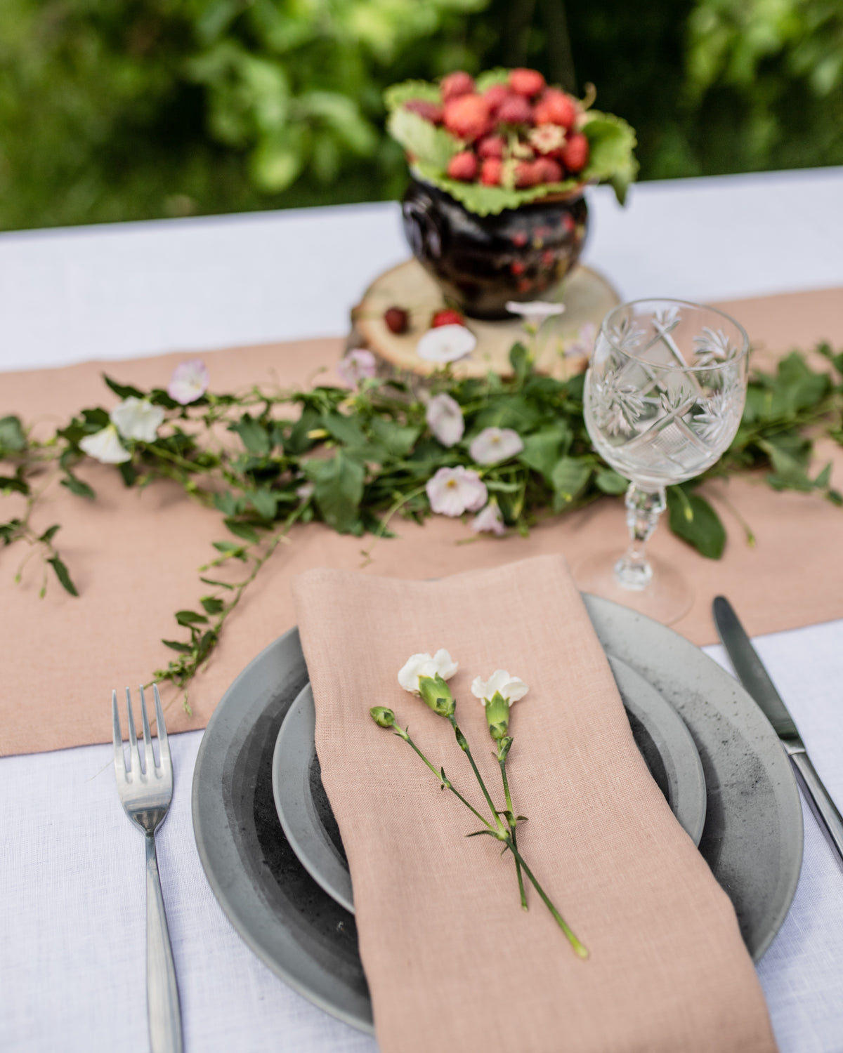 Elegant outdoor table setting with beige linen napkin, greenery, flowers, and cutlery.