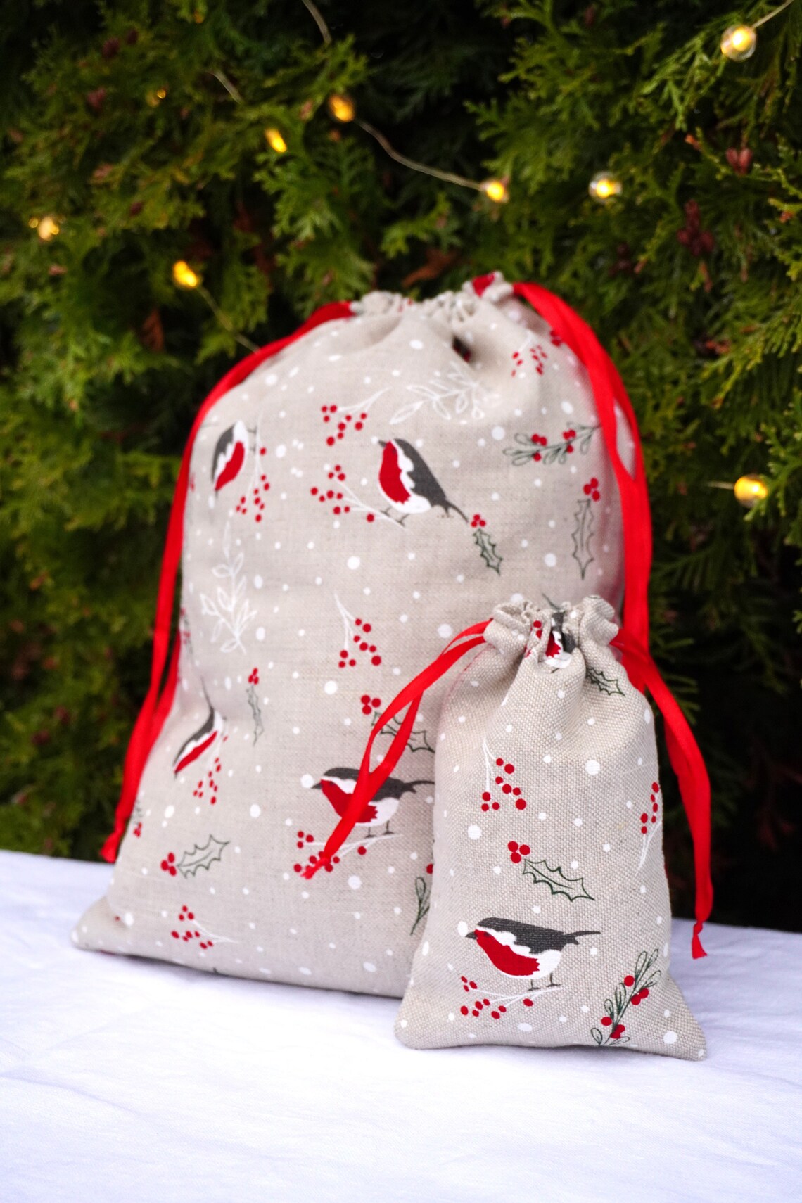 Two beige drawstring gift bags with a robin bird and holly berry print, accented with red drawstrings, displayed in front of a decorated Christmas tree.