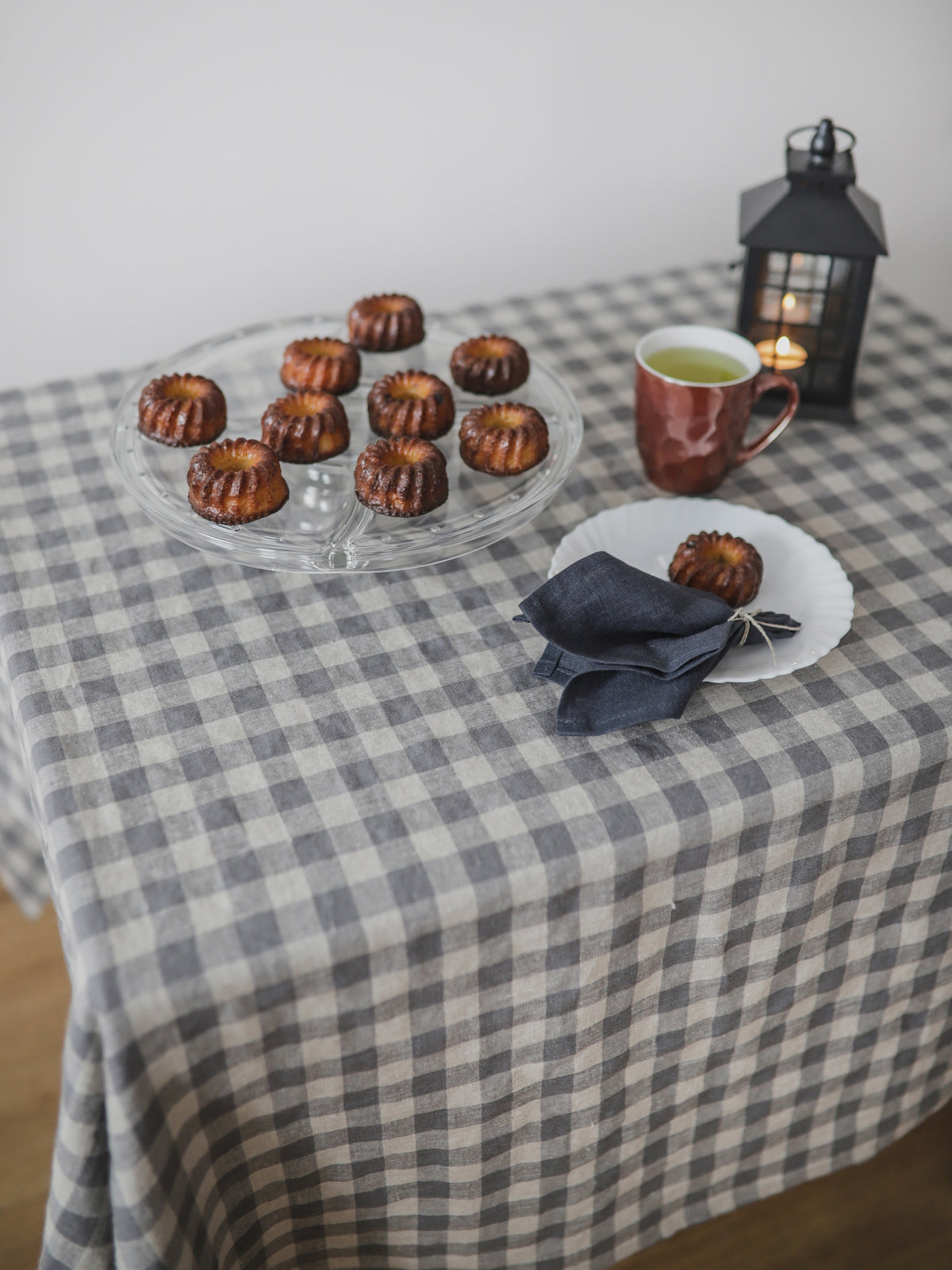 Table setting with small cakes, a mug of tea, and a candle on a checkered linen tablecloth.