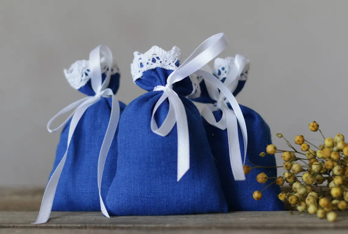 Three blue fabric pouches with white lace trim and white ribbons, arranged on a wooden surface with a small sprig of yellow flowers beside them against a neutral gray background.