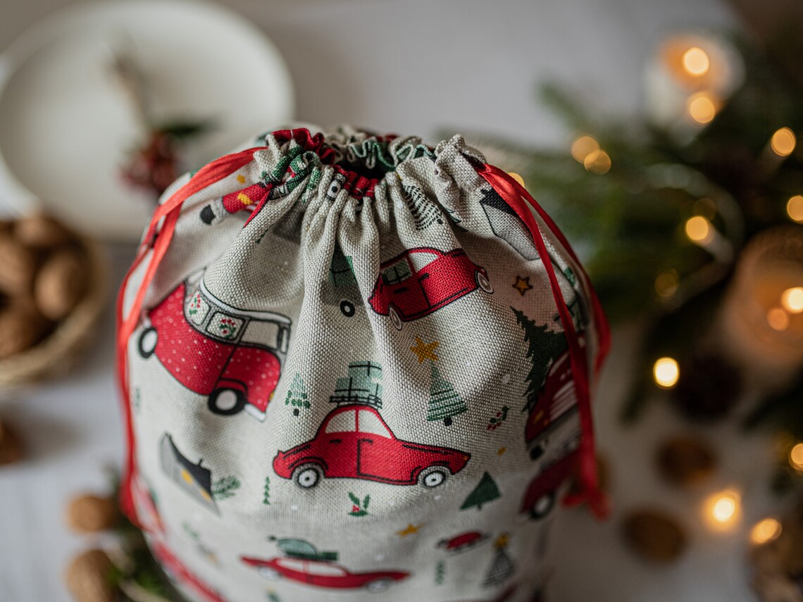 A linen fabric drawstring gift bag with a holiday print of red cars, green trees, and stars on a cream background, styled with festive bokeh lights and nuts in the background.