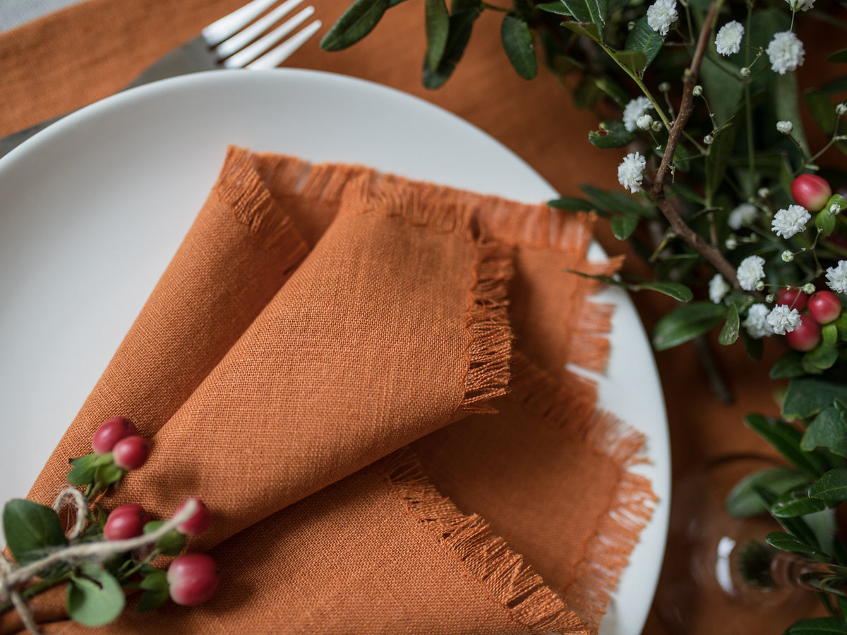 Fringed Orange linen napkin on a white plate with greenery and berries