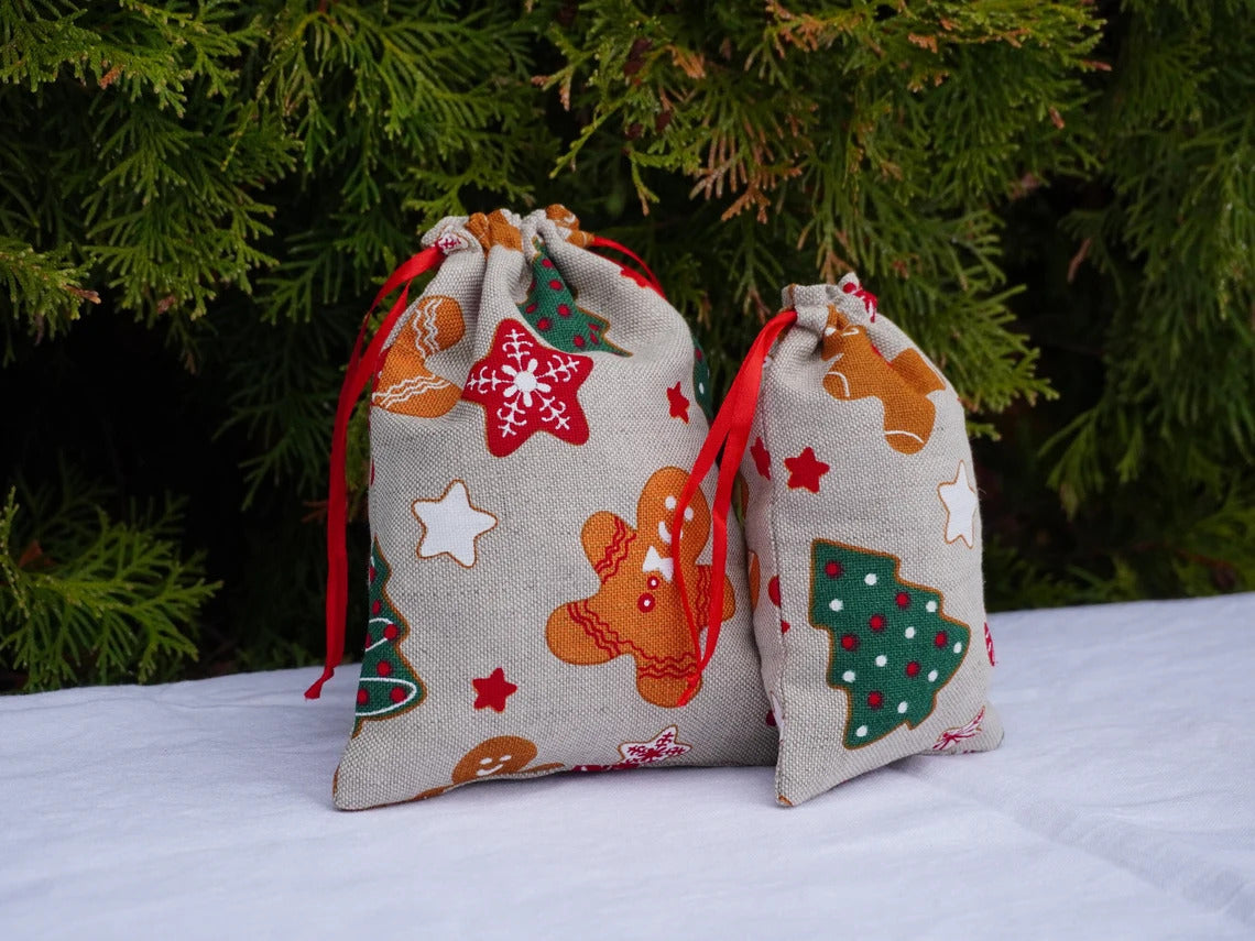 Two small linen drawstring gift bags with a festive Christmas print of gingerbread men, trees, and stars, set against a background of green pine branches.