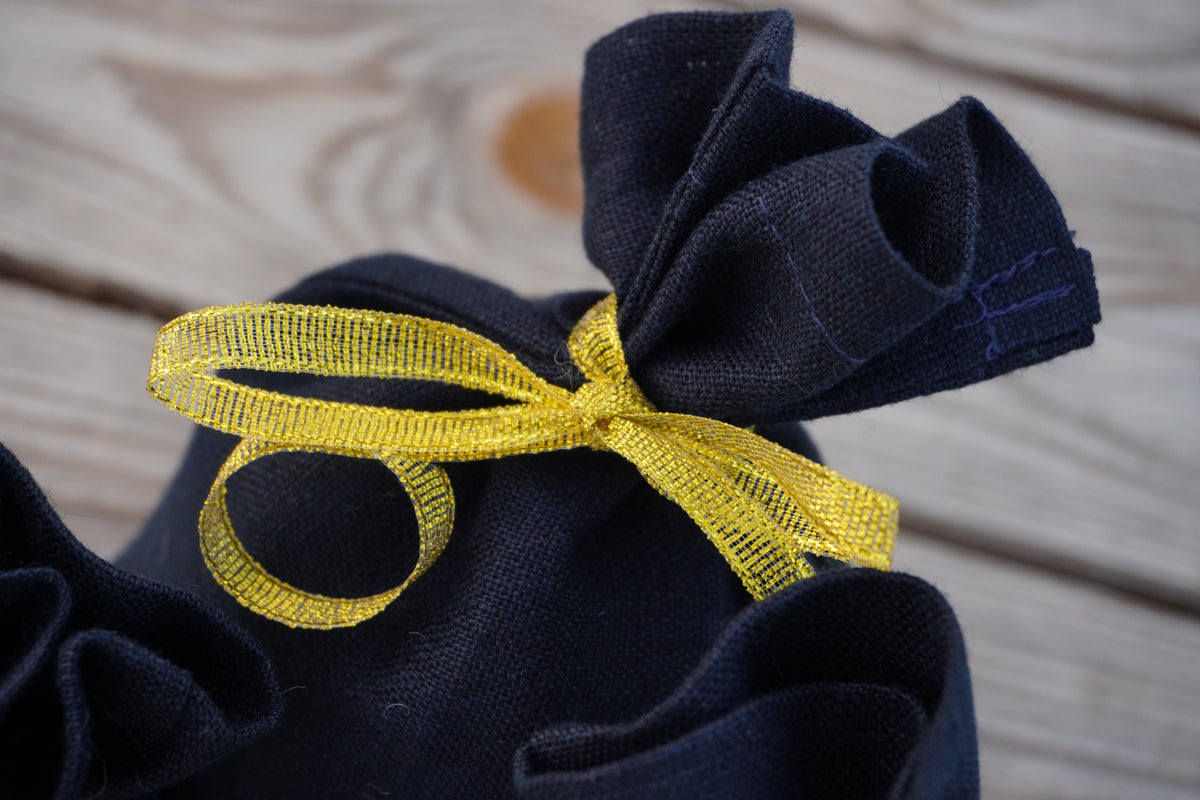 A close-up of a dark navy fabric pouch tied with a shiny yellow ribbon bow, set against a light wood plank background.