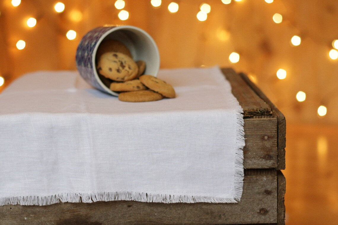 A not dyed fringed linen table runner draped over a rustic wooden crate, with a blue-and-white patterned cup tipped over to spill chocolate chip cookies. Warm bokeh string lights glow in the background.