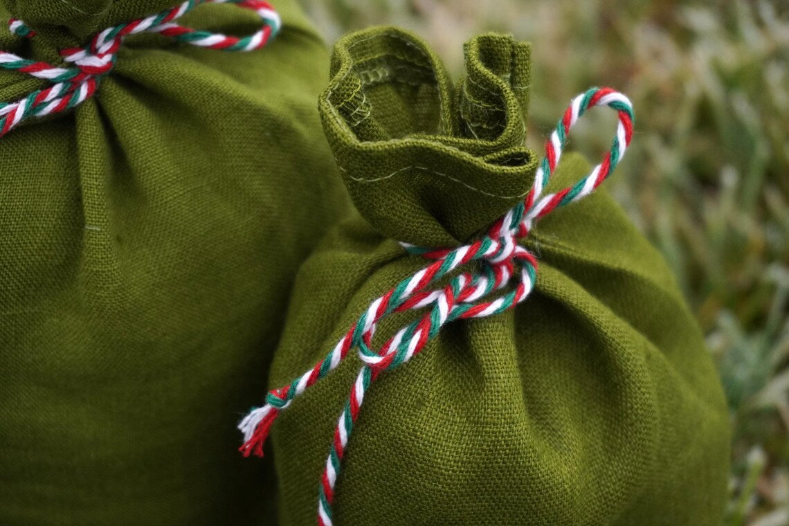 Two olive green linen bags tied with red, white, and green twisted cord, photographed outdoors against a blurred grassy background.