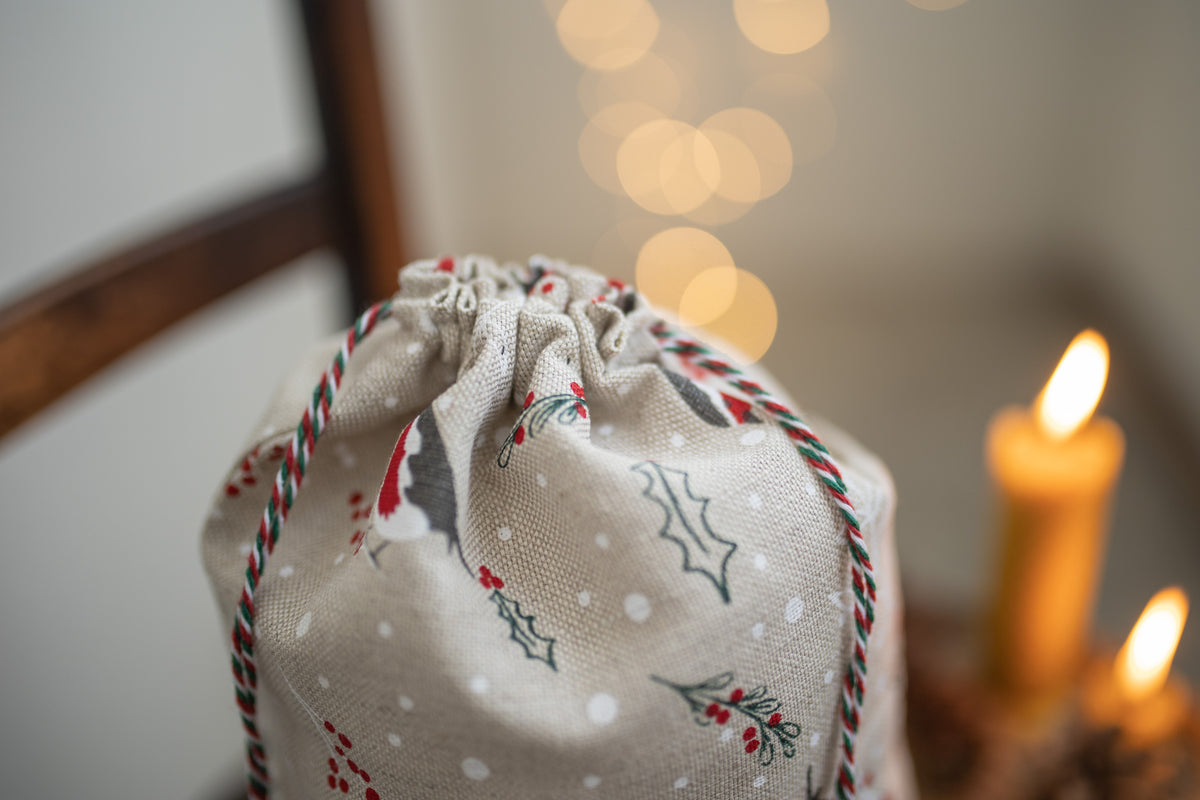 A close-up of a linen fabric Santa sack with a holiday print, featuring a beige base with white snowflakes, robin, green holly leaves, and red berries. The drawstring closure is finished with a red, green, and white striped braid.