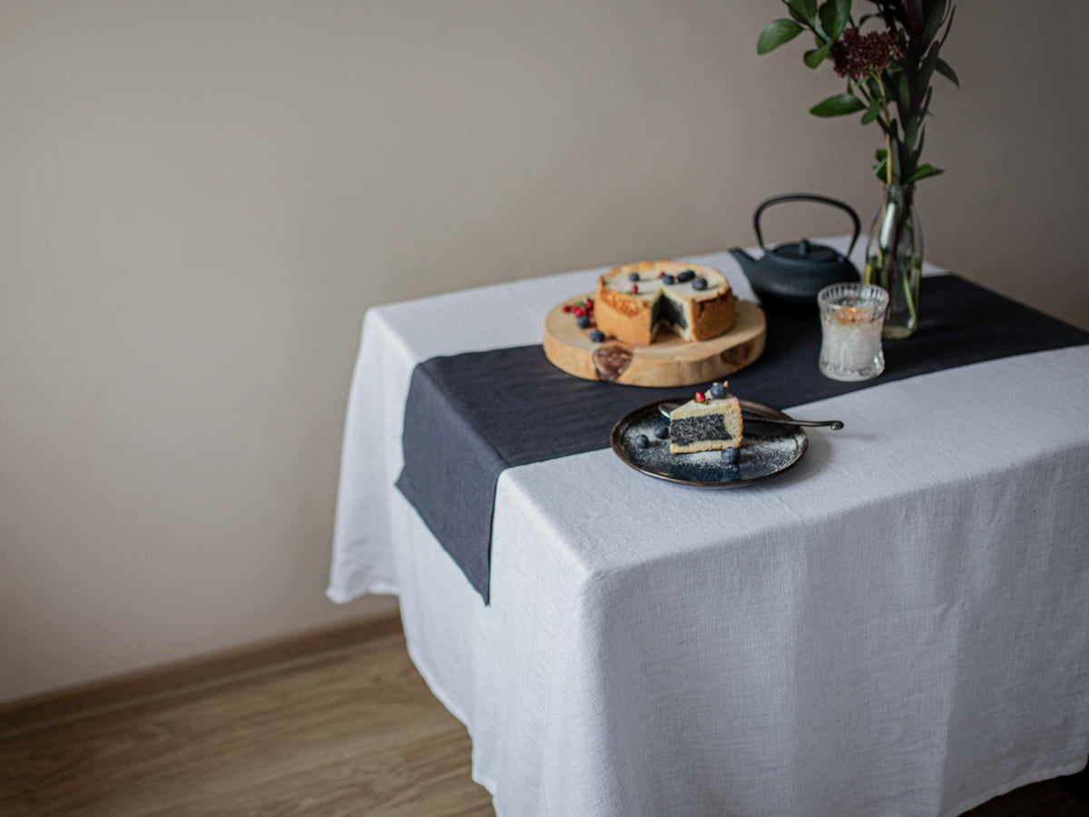 Table setting with a cake on a white linen tablecloth and a slice on a plate, against a plain wall.
