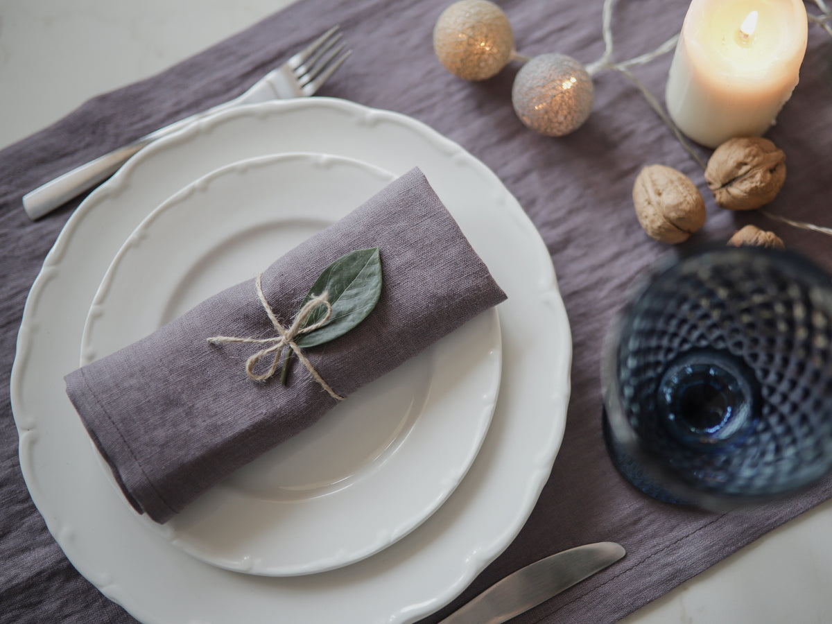 A styled table setting featuring a folded dusty lavender linen napkin in a muted dusty purple color, tied with twine and a small green leaf, placed on a white plate. The setting includes a matching purple runner, a blue textured glass, a white candle, and decorative walnuts.