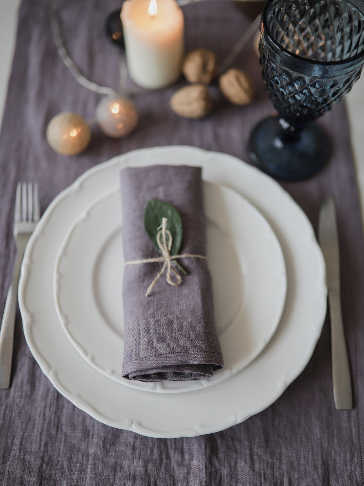 A styled table setting featuring a folded dusty purple linen napkin tied with twine and a small green leaf, placed on a stack of white ceramic plates. The setting includes a silver fork, knife, a dark blue textured goblet, walnuts, a lit candle, and decorative gold baubles on a purple tablecloth.