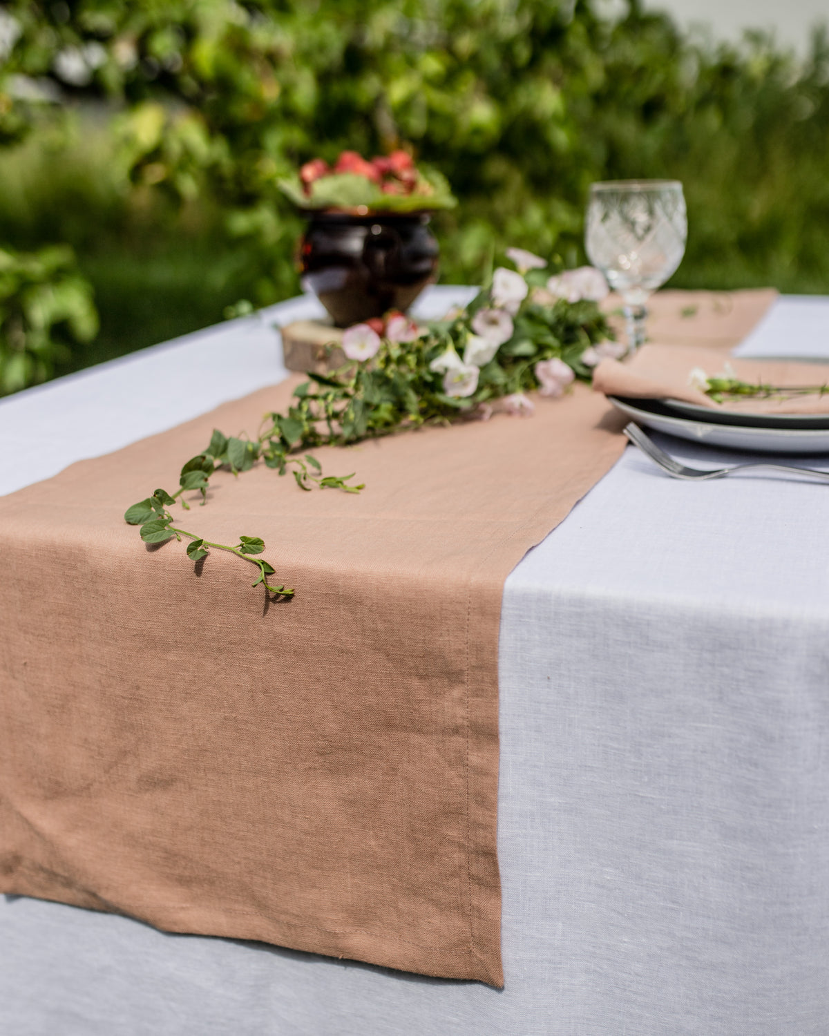 Table setting with a beige runner, white tablecloth, and floral centerpiece outdoors.