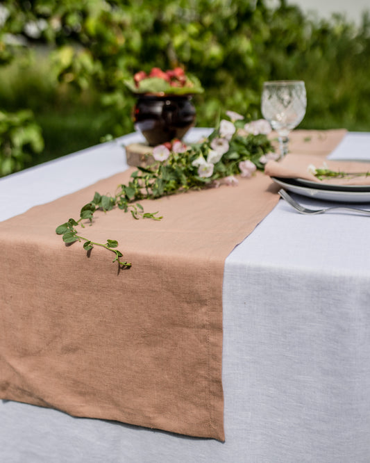 Table setting with a beige runner, white tablecloth, and floral centerpiece outdoors.