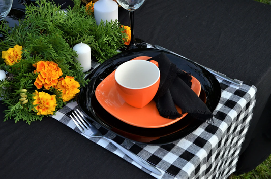 An outdoor table setting featuring a place setting with a bright orange ceramic cup and matching saucer, layered over black plates, a black napkin, a black-and-white checkered runner, and a green and orange floral centerpiece.
