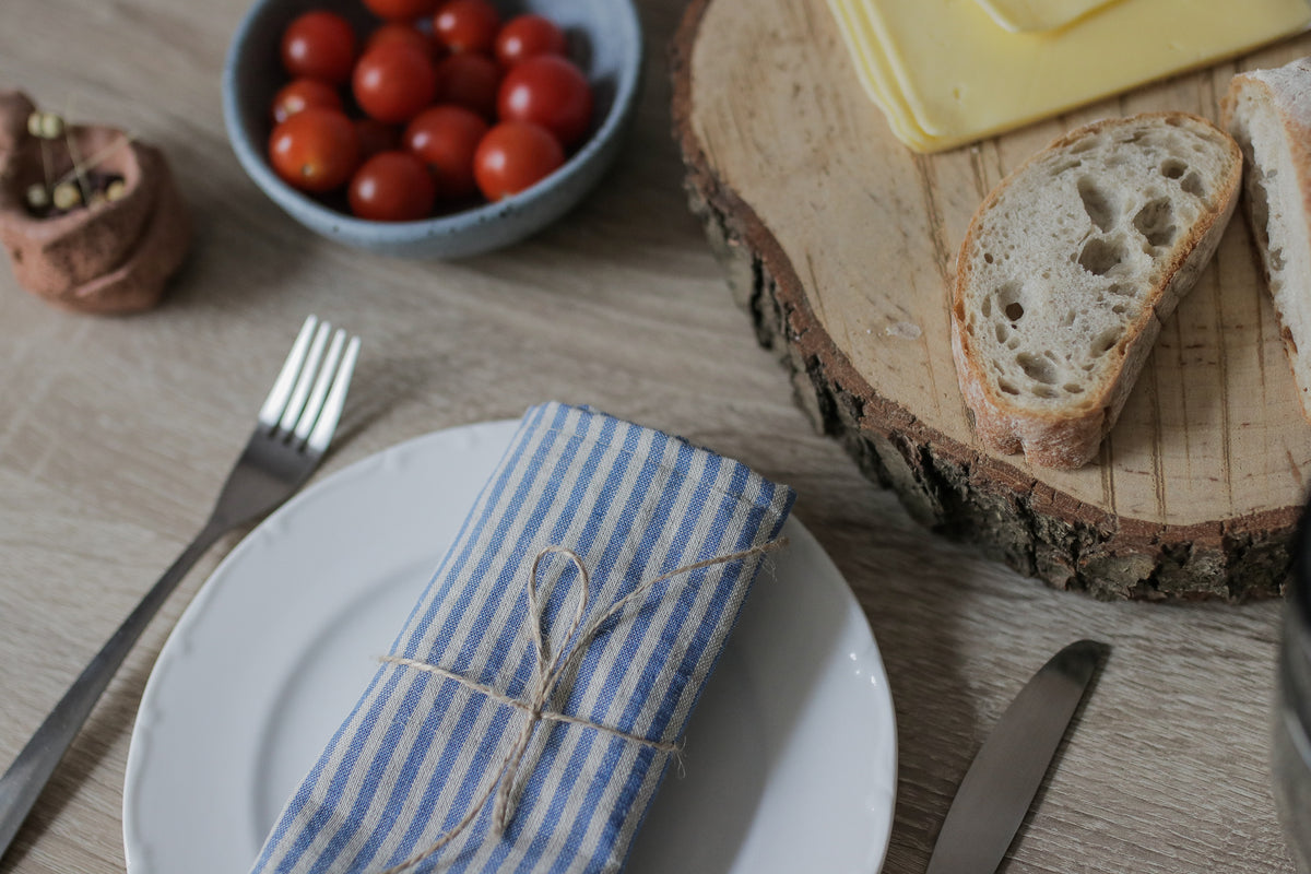 Table setting with bread, tomatoes, and a blue striped linen napkin on a wooden surface.