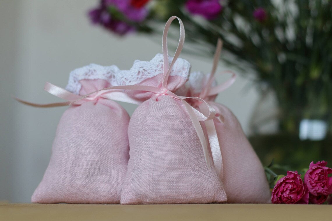 Three light pink linen fabric pouches with white lace trim and pink ribbons, arranged on a wooden surface with blurred flowers in the background.