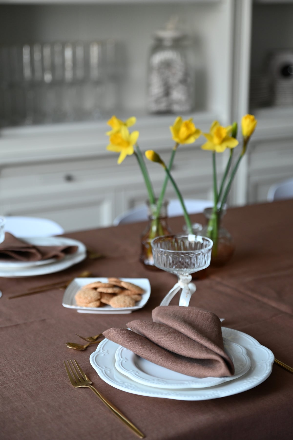 Dining table setting with brown linen napkins, white plates, and yellow flowers in the background.