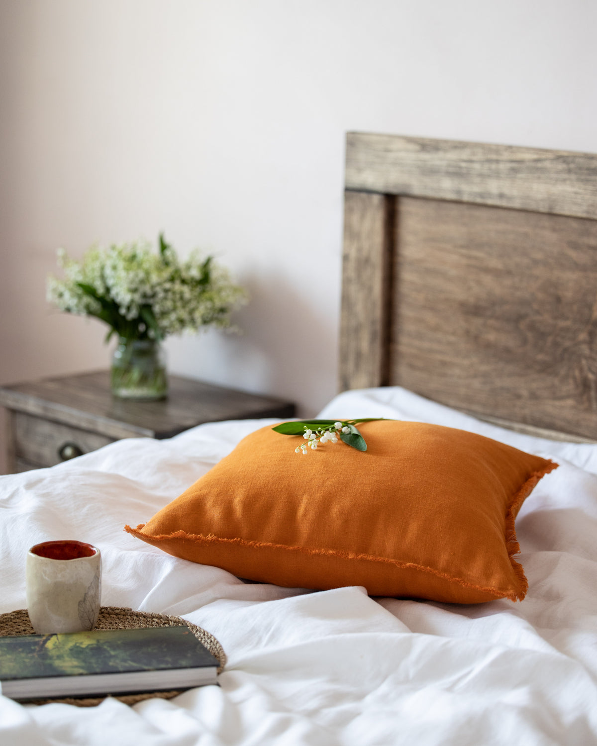 Orange pillow on a bed with a wooden headboard and a vase of flowers on a nightstand.