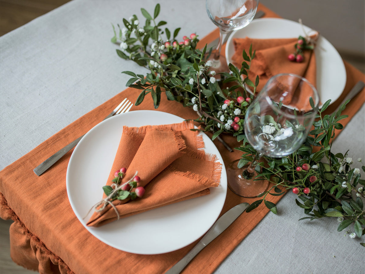 Table setting with burnt orange fringed  napkins, white plates, and greenery on a wooden table.