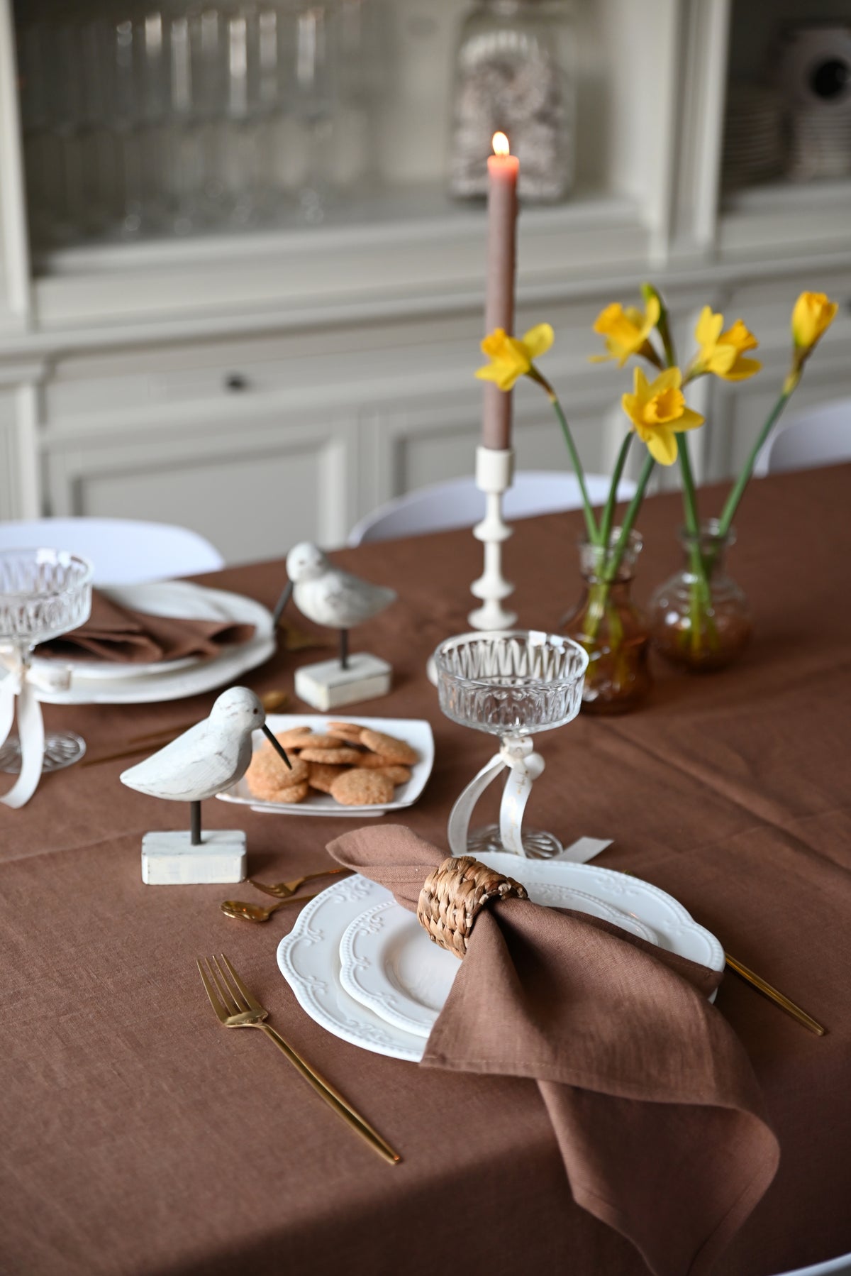 Decorative table setting with candles, flowers, and place settings on a brown tablecloth with brown linen napkins.