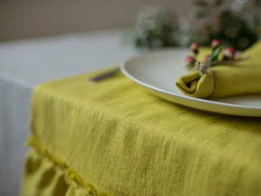 A close-up of a chartreuse linen tablecloth with a ruffled hem, styled with a white plate and a matching chartreuse napkin on a dining table.