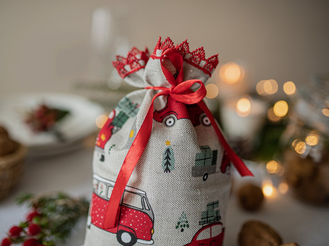 A linen  fabric gift bag with a red and green holiday print of vintage cars and Christmas trees, tied with a red ribbon and topped with a red lace crown, set against a warm, softly blurred background with bokeh lights.