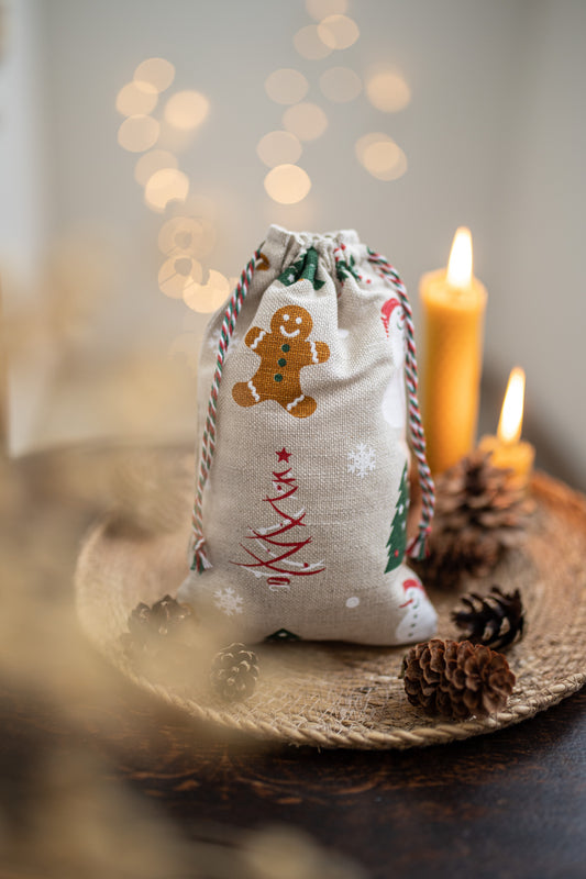 A small linen fabric Christmas gift bag with a drawstring closure, printed with a gingerbread man, Christmas tree, and snowflakes, styled on a woven tray with lit candles and pinecones.