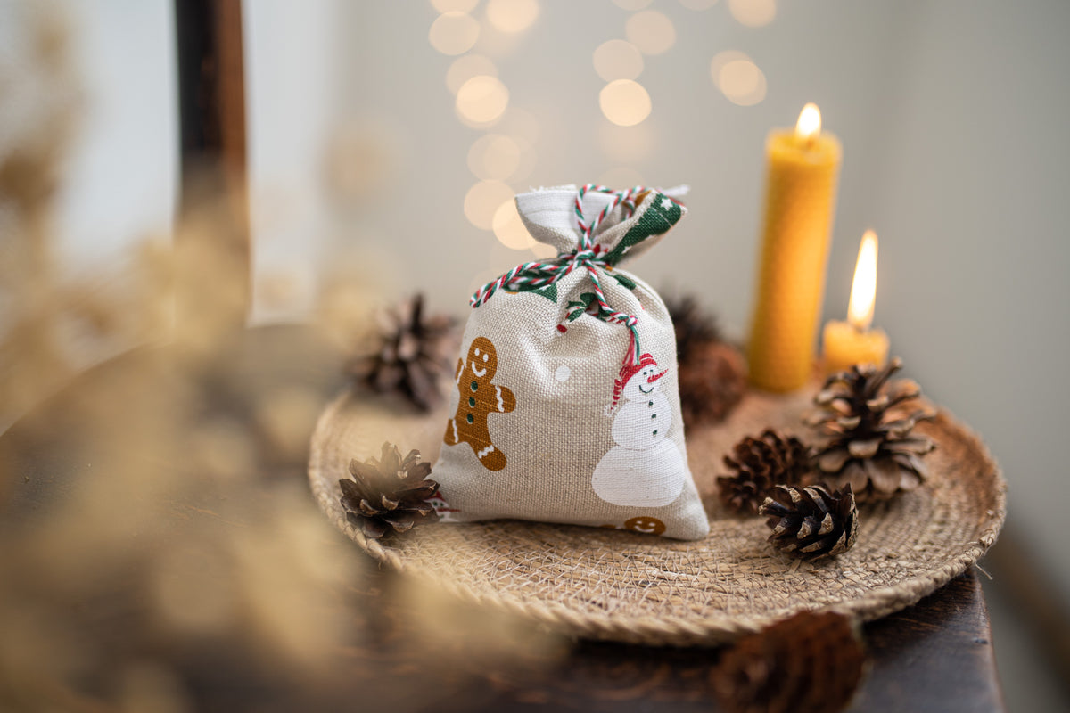 A small linen fabric Christmas gift bag with gingerbread and snowman prints sits on a woven tray surrounded by pinecones, with lit candles and warm bokeh lights in the background.