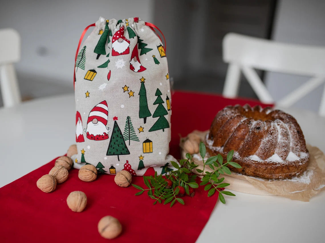 A festive Christmas-themed fabric drawstring bag with a gnome and tree print, styled on a red table runner next to a bundt cake, walnuts, and greenery.