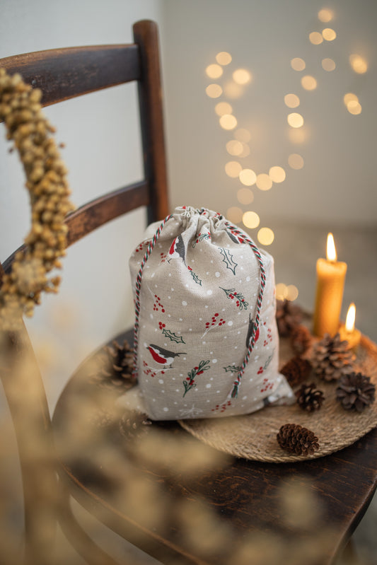 A small drawstring gift bag with a Christmas bird and holly pattern sits on a rustic wooden chair surrounded by candles, pinecones, and warm bokeh lights in the background.