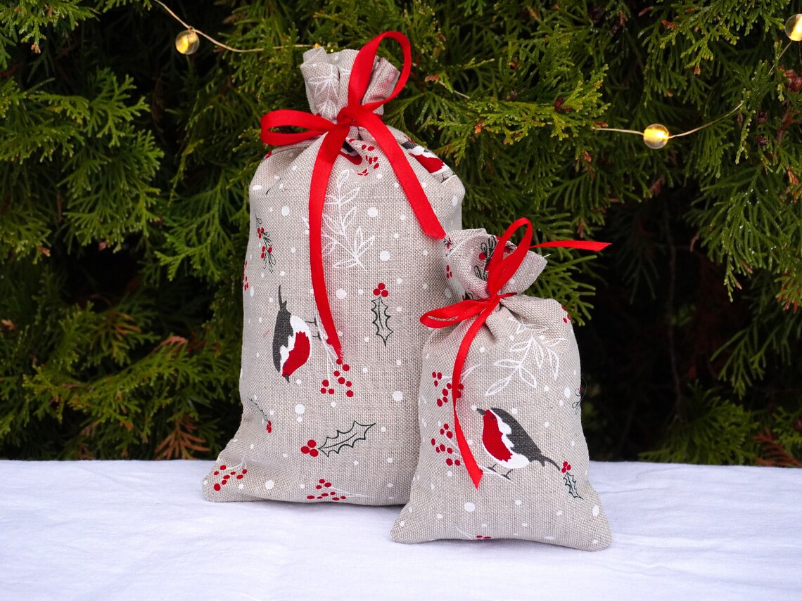 Two linen fabric drawstring gift bags in a light beige holiday print with red bird motifs, red berries, and white snowflakes, tied with red ribbon bows, set against a dark green pine tree background with warm string lights.
