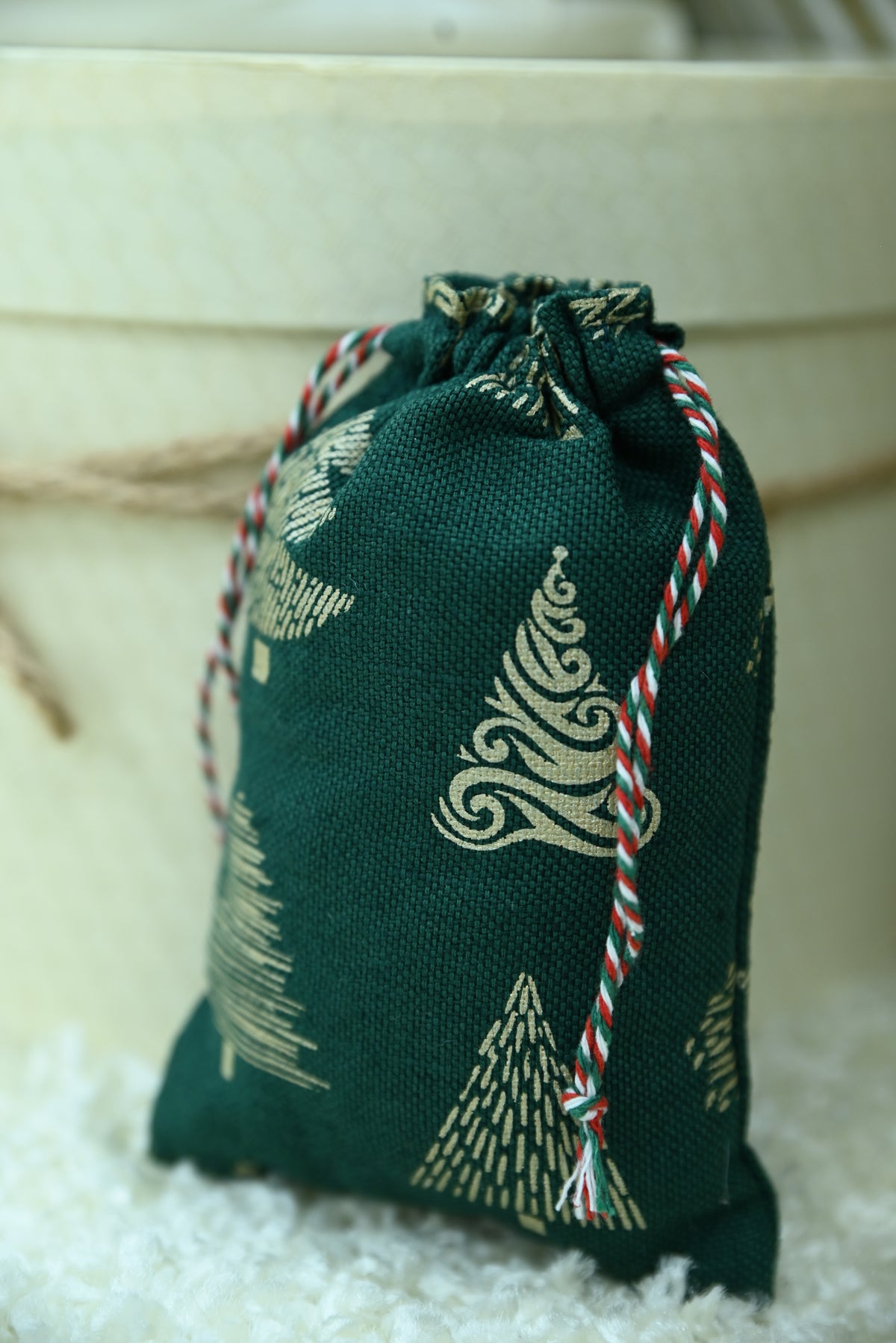 A small dark green drawstring pouch with a cream Christmas tree pattern, resting on a white textured surface in front of a beige background.
