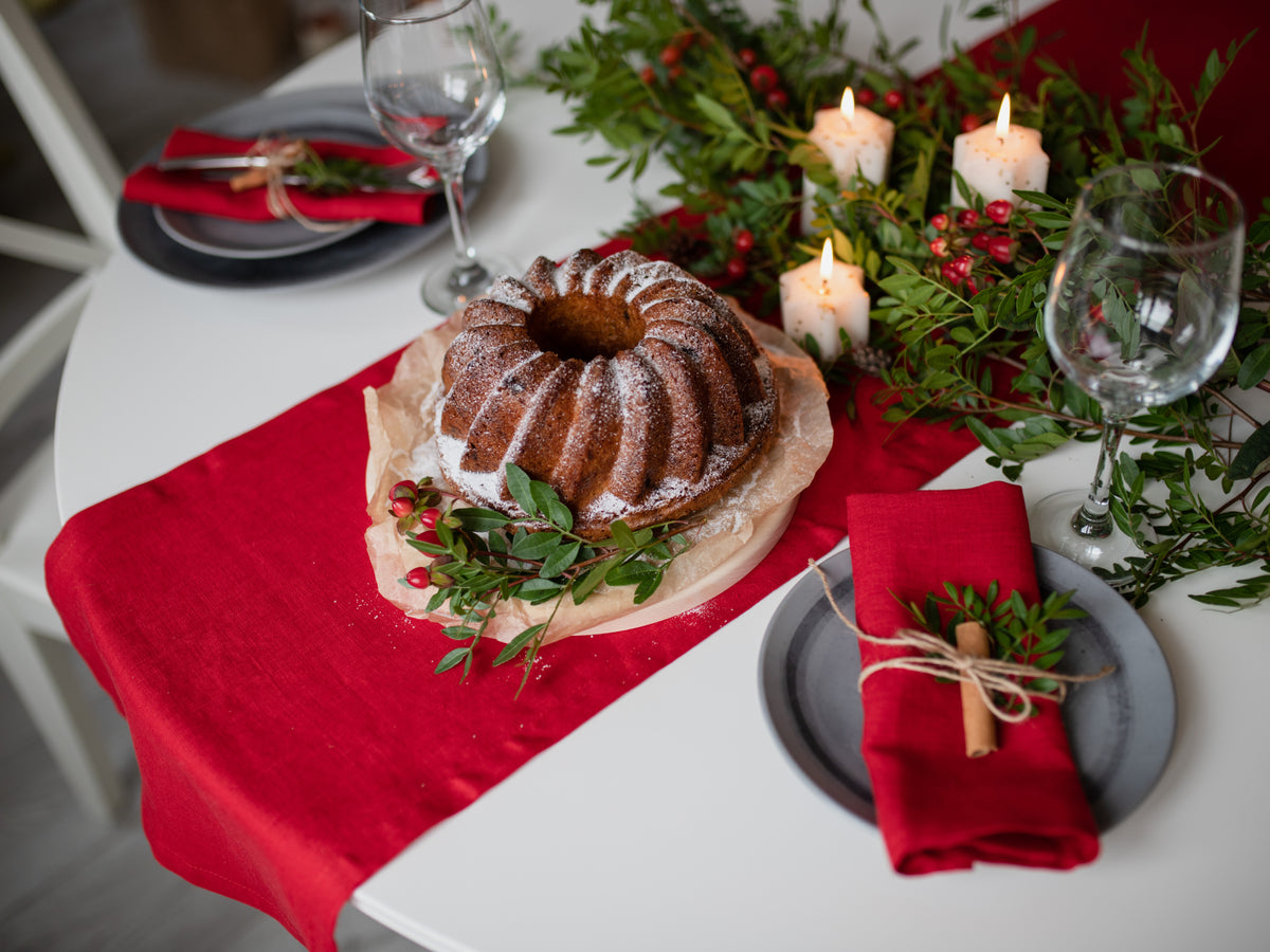 A holiday-themed table setting featuring a ring-shaped bundt cake dusted with powdered sugar on parchment paper, placed on a red velvet runner. The table is decorated with green pine branches, red berries, white pillar candles, and place settings with red linen  napkins and gray plates.