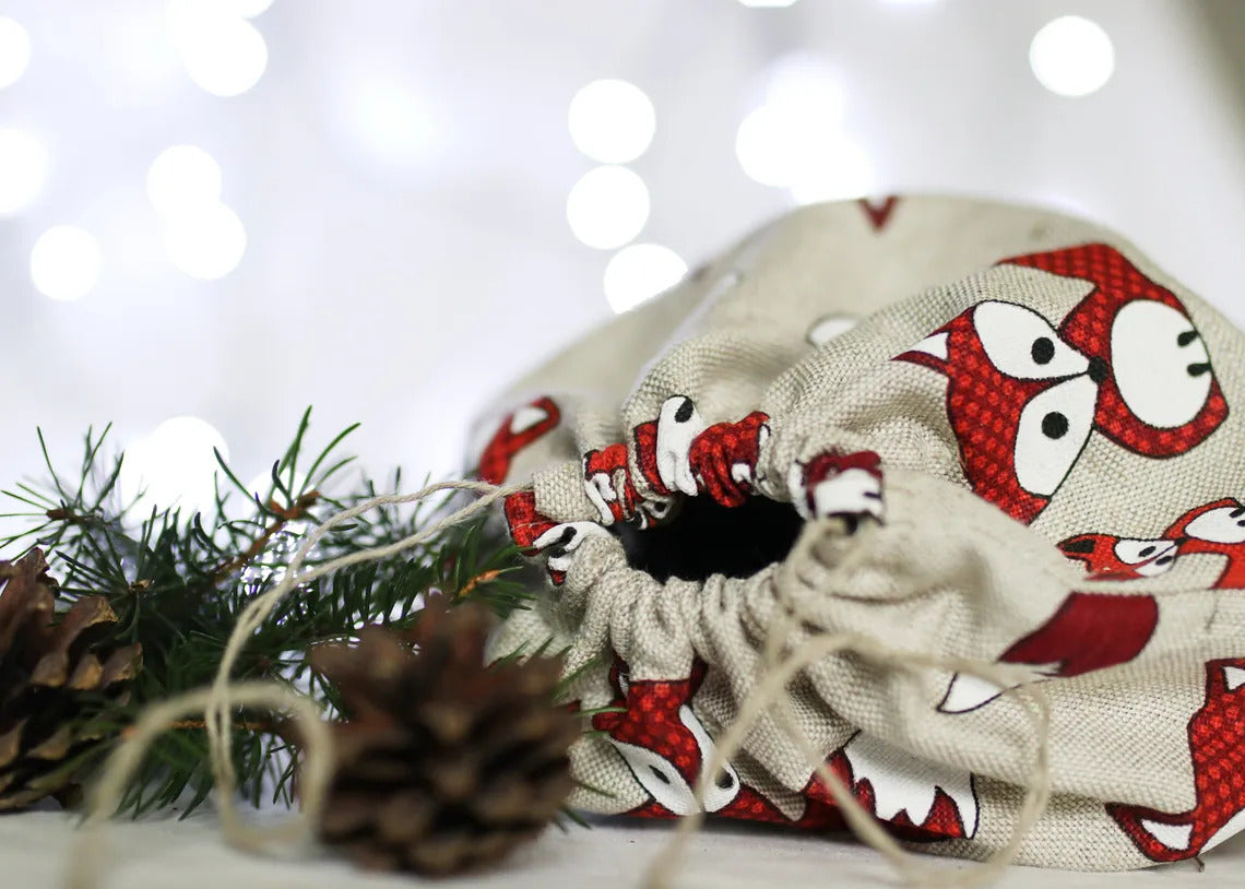 A fabric drawstring gift bag   fox pattern, styled with pine needles and pinecones against a soft white bokeh background.