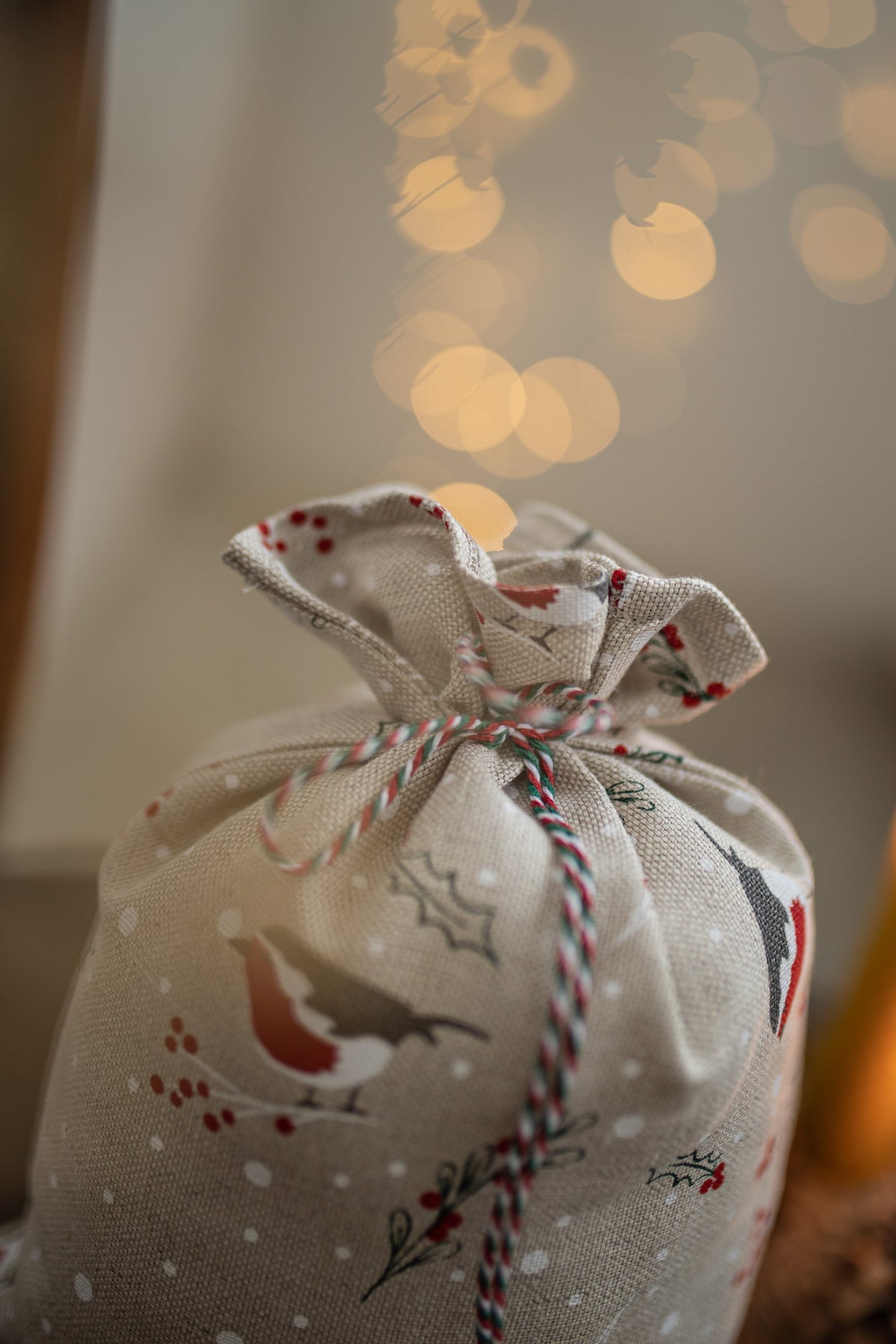 A gift bag made of natural linen with a Christmas bird and holly print, tied with red, white, and blue striped twine, set against a warm background with soft bokeh lights.