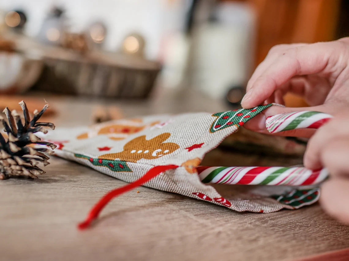 A close-up of hands placing a red and white striped candy cane into a small fabric holiday pouch with gingerbread and Christmas patterns, on a wooden tabletop.