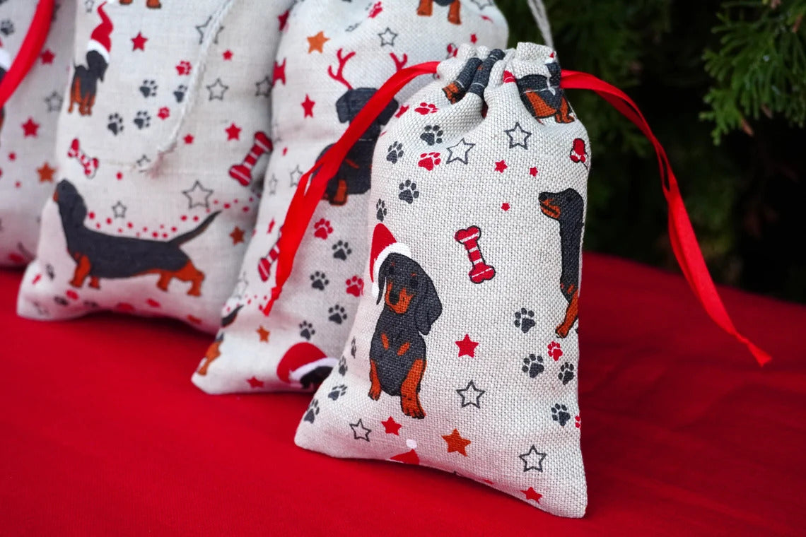 Three small linen fabric drawstring gift bags with a dachshund print, red ribbon ties, and holiday-themed motifs like stars, paw prints, and a bone, arranged on a red surface with a blurred green background.
