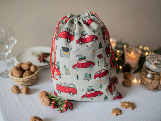 A linen fabric drawstring gift bag with a red vintage car and Christmas tree print, styled on a table with cookies, walnuts, and candles.