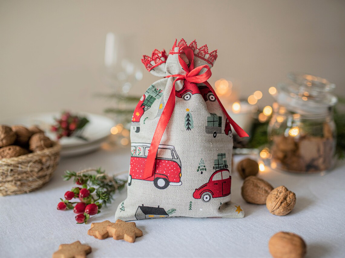A small linen fabric gift bag with a red and green holiday print of vans and cars, tied with a red ribbon, set on a table with cookies, walnuts, and warm bokeh lights in the background.
