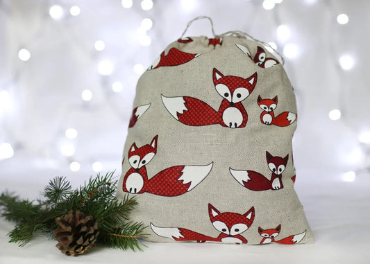 A linen fabric drawstring bag with a repeating red and white fox print on a light beige linen-like background, styled with pine branches and a pinecone against a soft white bokeh background.