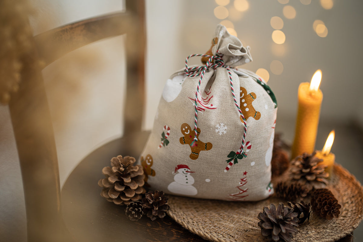 A small fabric gift bag with a gingerbread man and snowman print, tied with a striped string, displayed on a table with pinecones and lit candles.