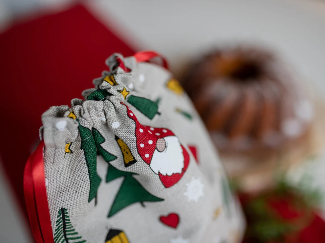 A close-up of a fabric drawstring pouch with a holiday gnome and Christmas tree print, tied with a red ribbon
