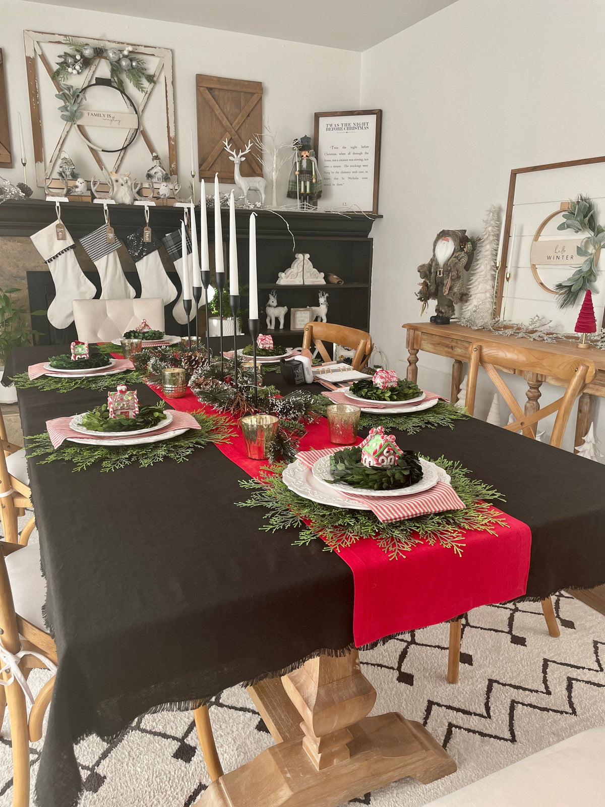 A styled holiday dining table in a home setting, featuring a black tablecloth with a bright red linen runner, white plates, red striped napkins