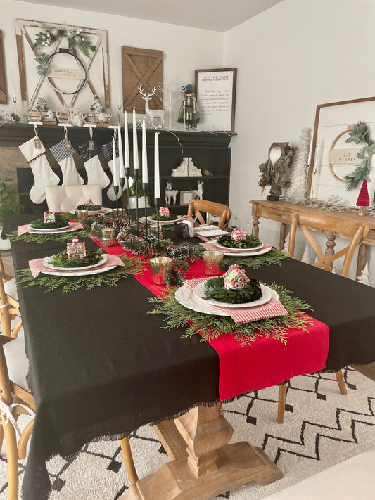 A styled holiday dining table in a home setting, featuring a black tablecloth with a bright red linen runner, white plates, red striped napkins