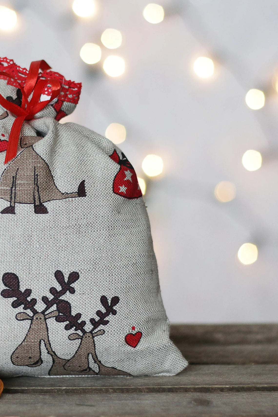 A rustic linen sack with a red ribbon closure, decorated with cartoon reindeer and small red hearts, set on a wooden surface with warm bokeh string lights in the background.