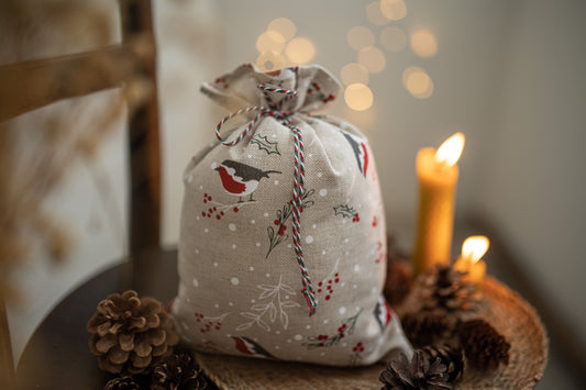 A linen gift bag with a winter bird and holly print, tied with red-and-white striped twine, styled on a woven tray with pinecones and lit candles in a warm indoor setting.