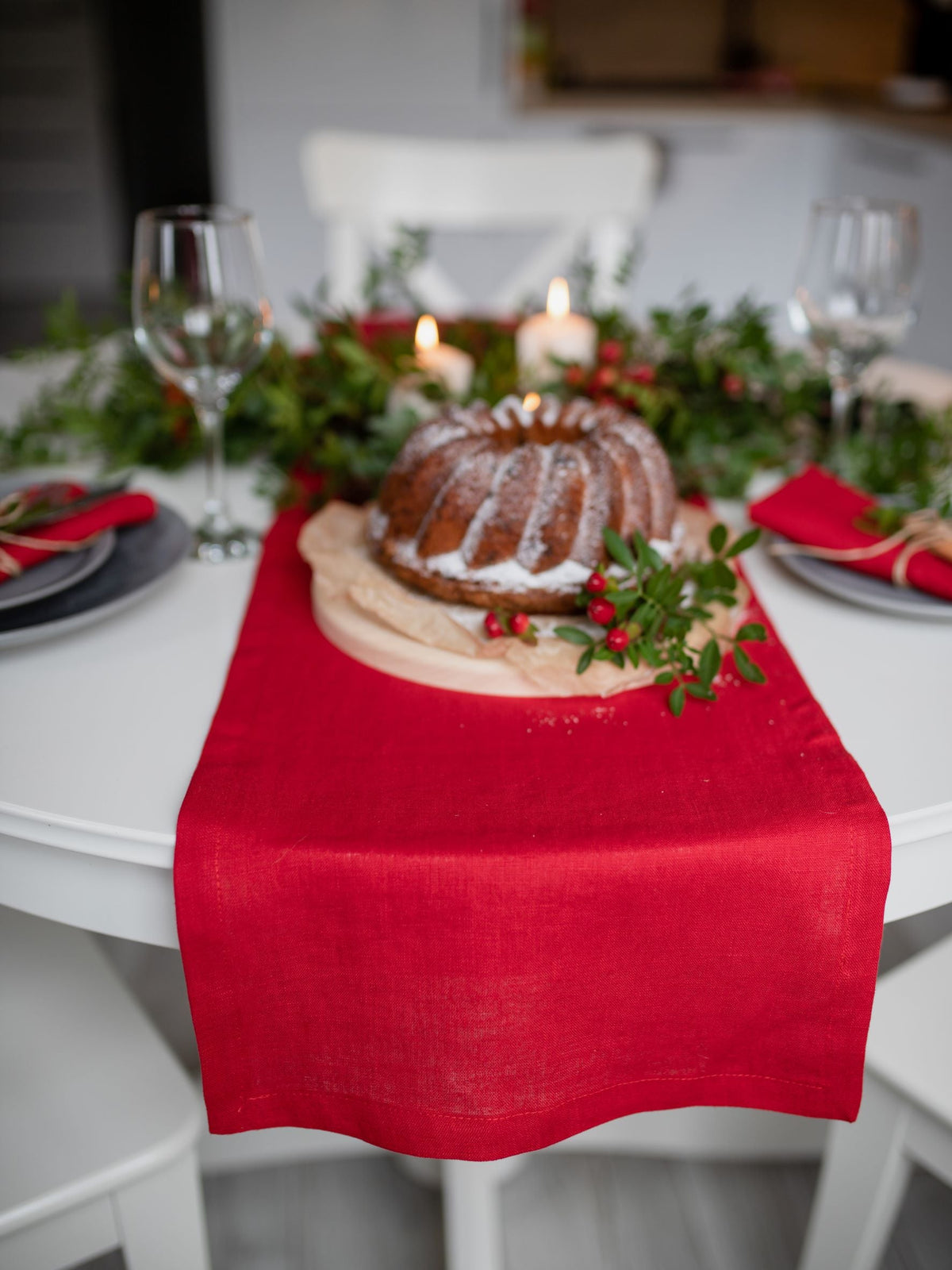 A festive holiday table setting featuring a red linen table runner draped over a white table, styled with a decorated bundt cake, green garlands, candles, and wine glasses.