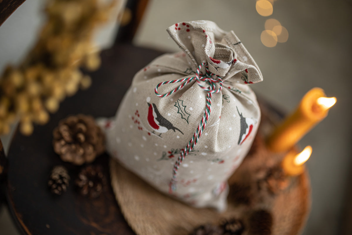 A linen fabric Santa sack with a Christmas bird pattern, tied with a red and white striped twine bow, styled on a rustic wooden surface with pinecones and lit candles.