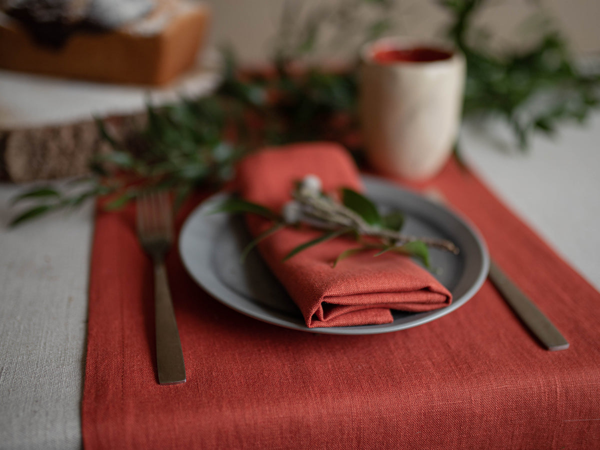 A styled table setting featuring a clay linen table runner, a folded clay napkin on a gray plate, silver flatware, and greenery in a softly blurred rustic setting.