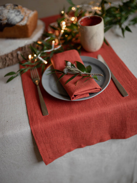 A styled table setting featuring a rust-orange (clay) linen table runner, a matching folded napkin on a gray plate, gold-toned cutlery, greenery, and warm string lights in a cozy indoor setting.