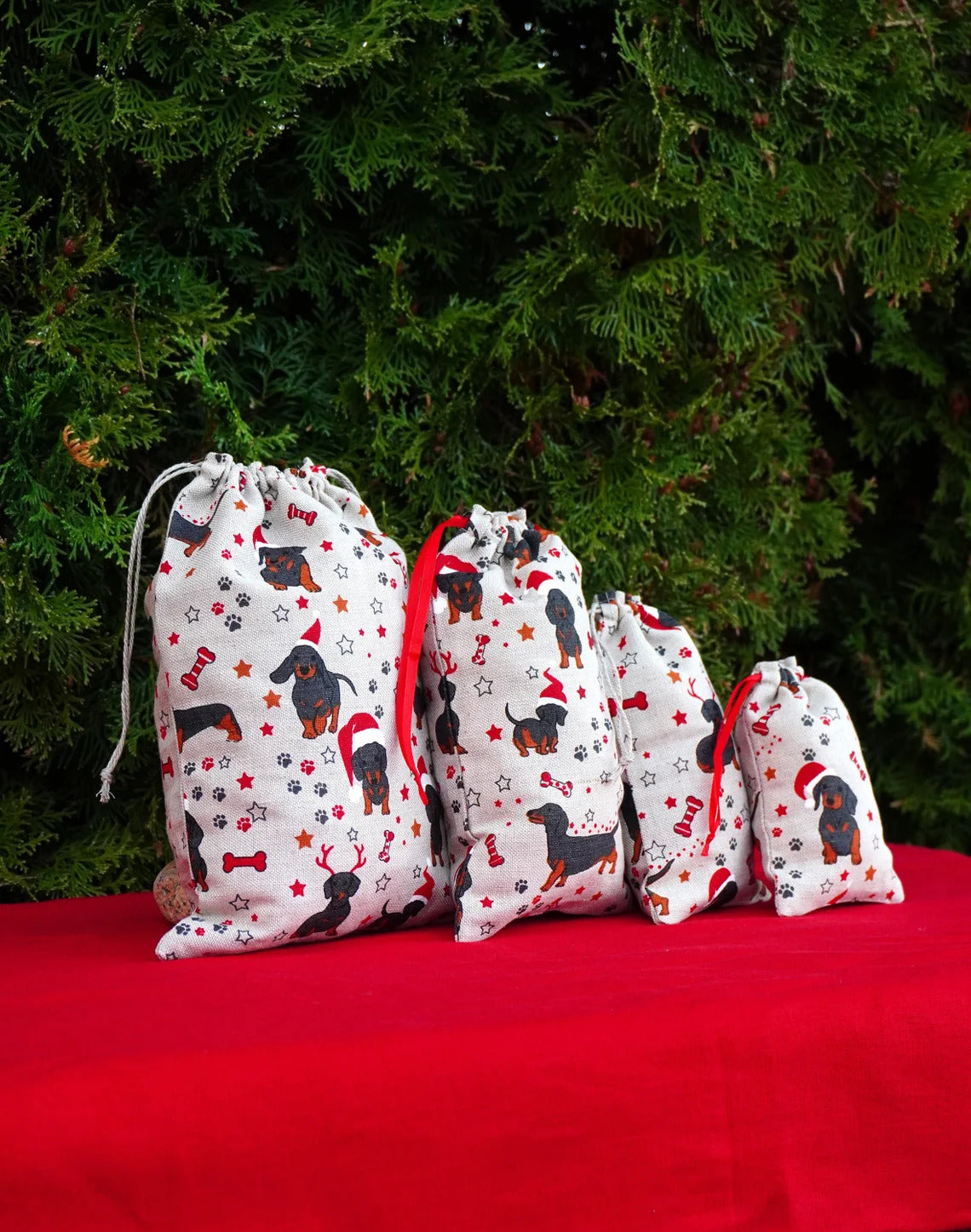 Four drawstring gift bags in descending sizes, printed with black dachshund dogs wearing red Santa hats and surrounded by red stars and bones on a white background, set against a red fabric surface and a green pine background.